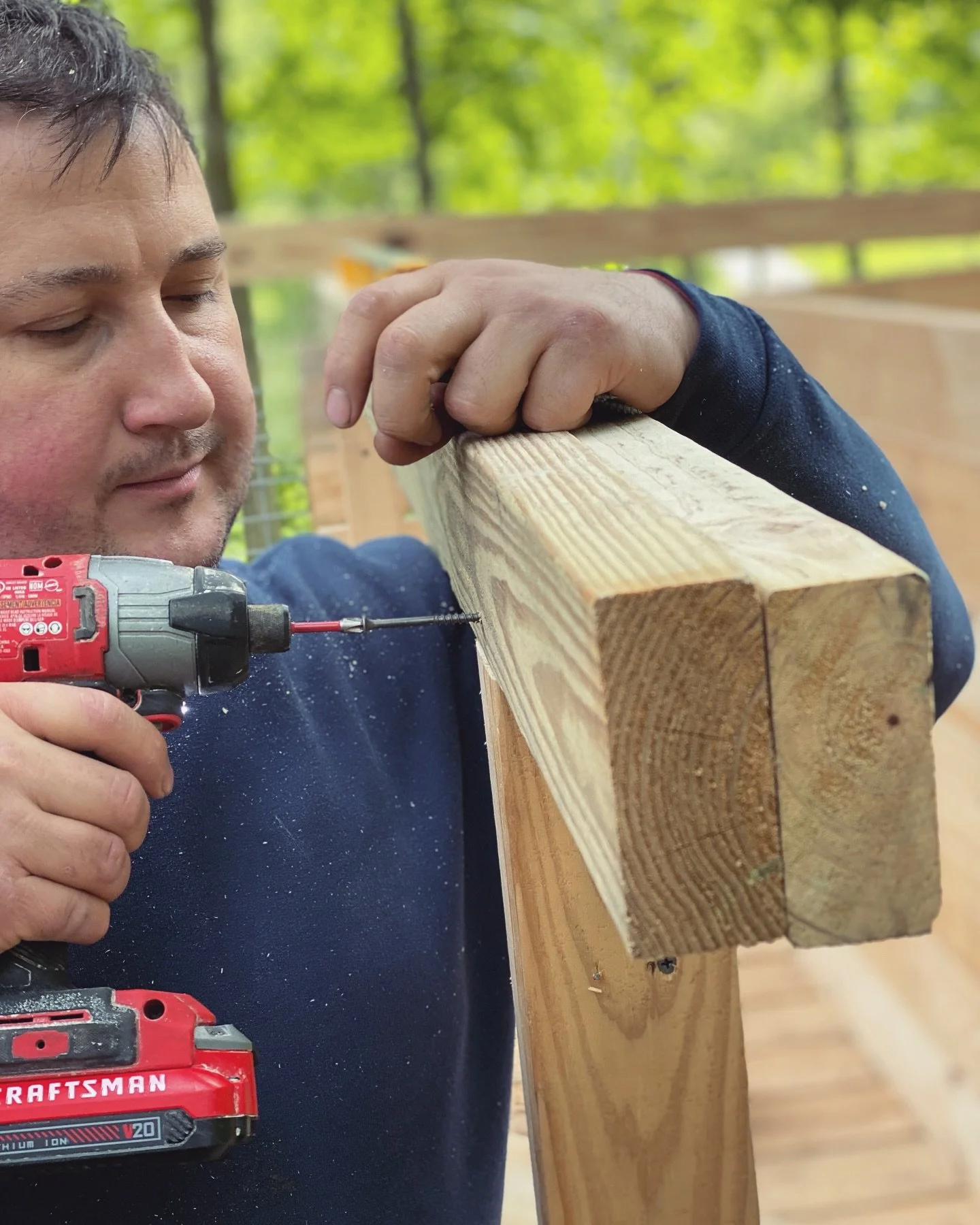 Man drilling into a wooden beam outdoors, with green trees in the background.