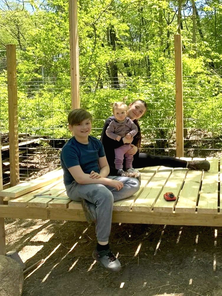 Three children sitting on a wooden platform in a wooded outdoor setting. Girl is sitting with one leg extended and holding a baby, while a boy sits cross-legged nearby. There are wooden posts, wire fencing, and trees in the background.