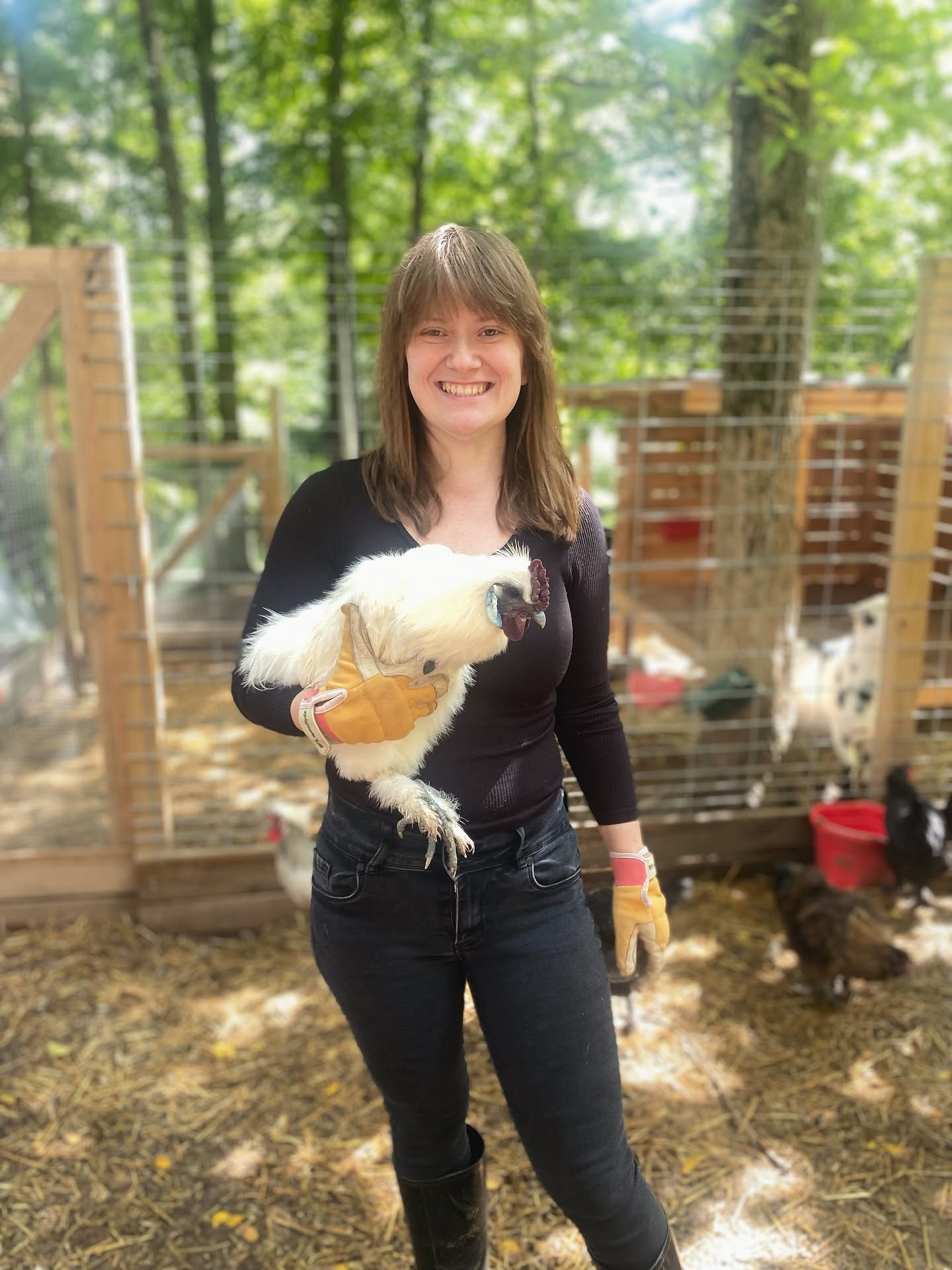 Woman smiling while holding a white chicken outdoors with a wooden and wire enclosure in the background.