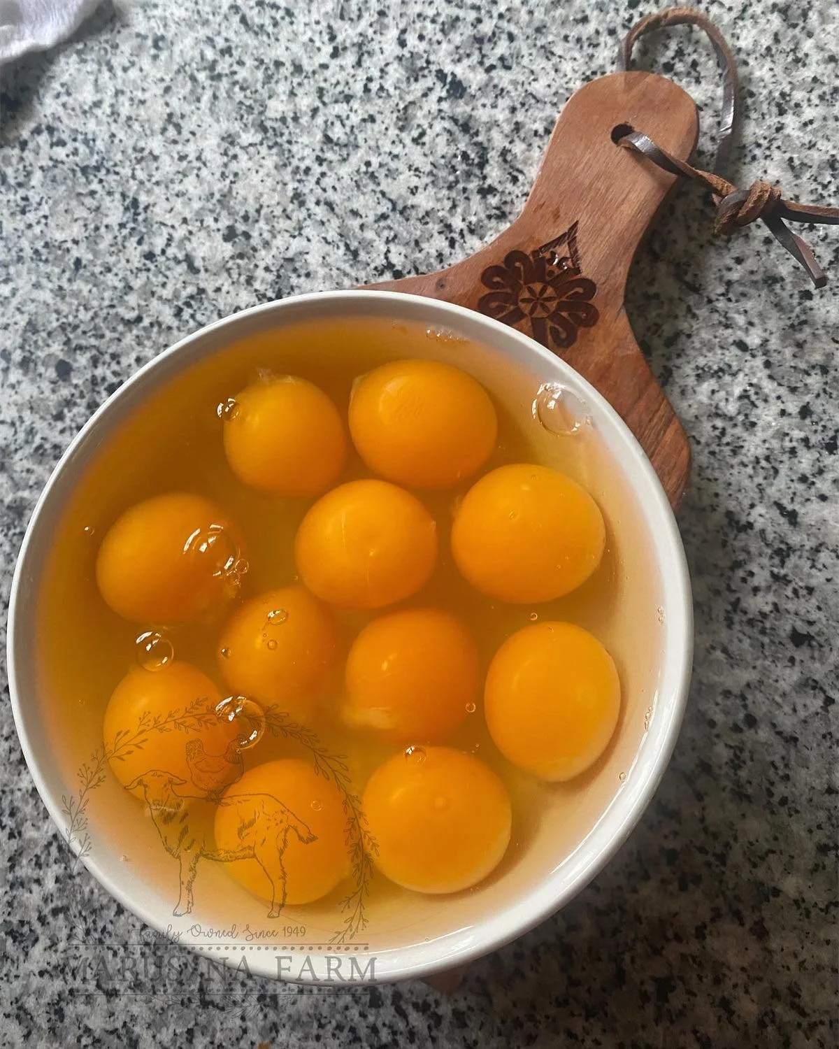 A bowl of cracked eggs on a speckled gray countertop, with a wooden cutting board