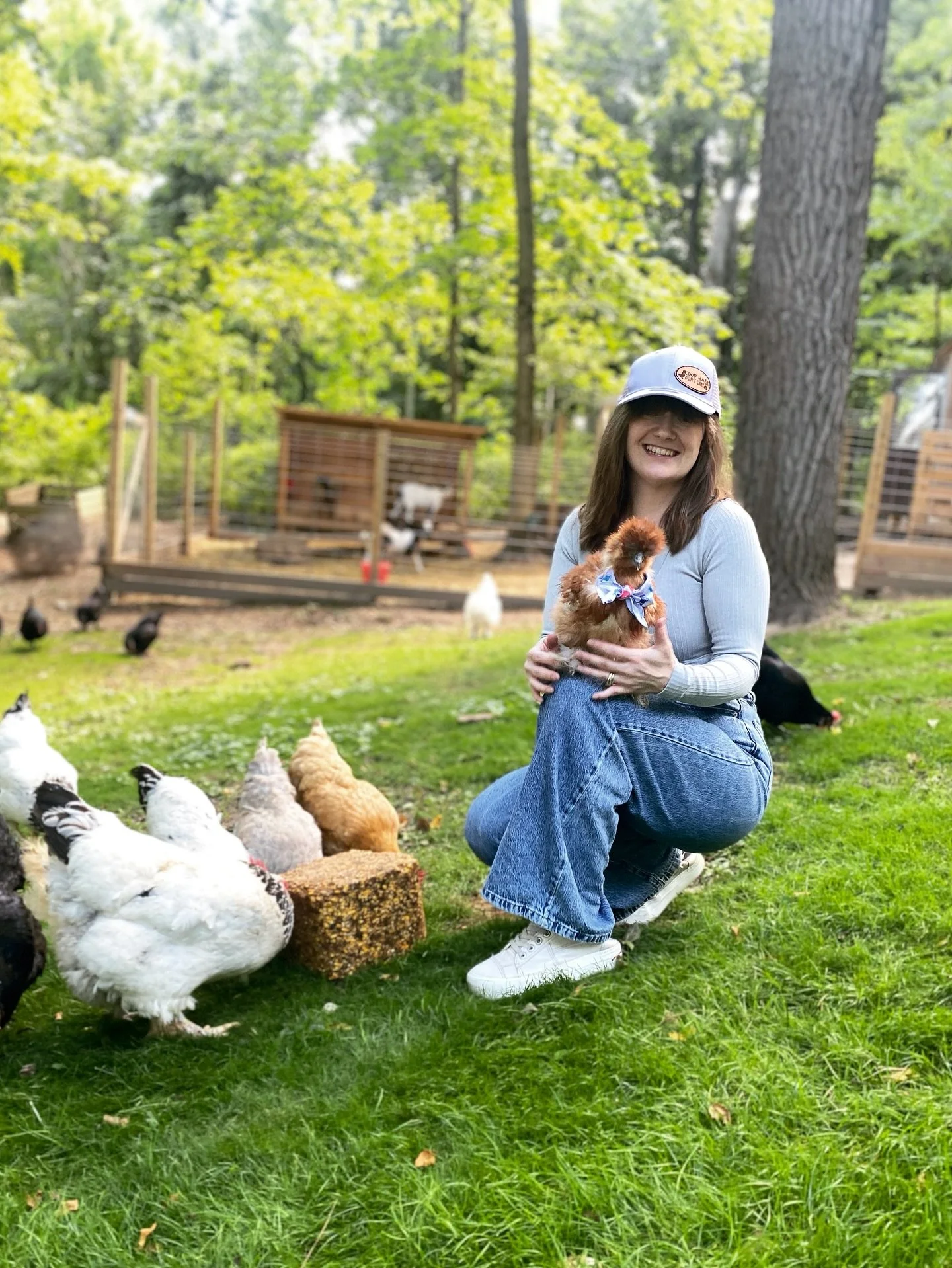 A woman kneeling on yellow grass in a park-like setting, holding a small, fluffy, brown chicken wearing a bowtie, with other chickens and ducks around her and chickens in cages in the background.