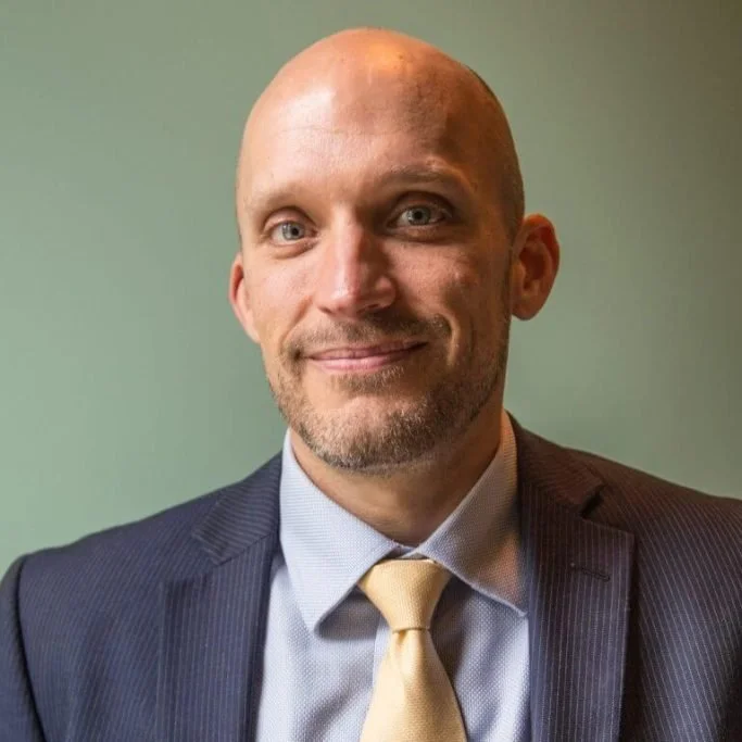 A man with a bald head and facial hair wearing a dark suit, light dress shirt, and yellow tie, standing against a plain green wall.