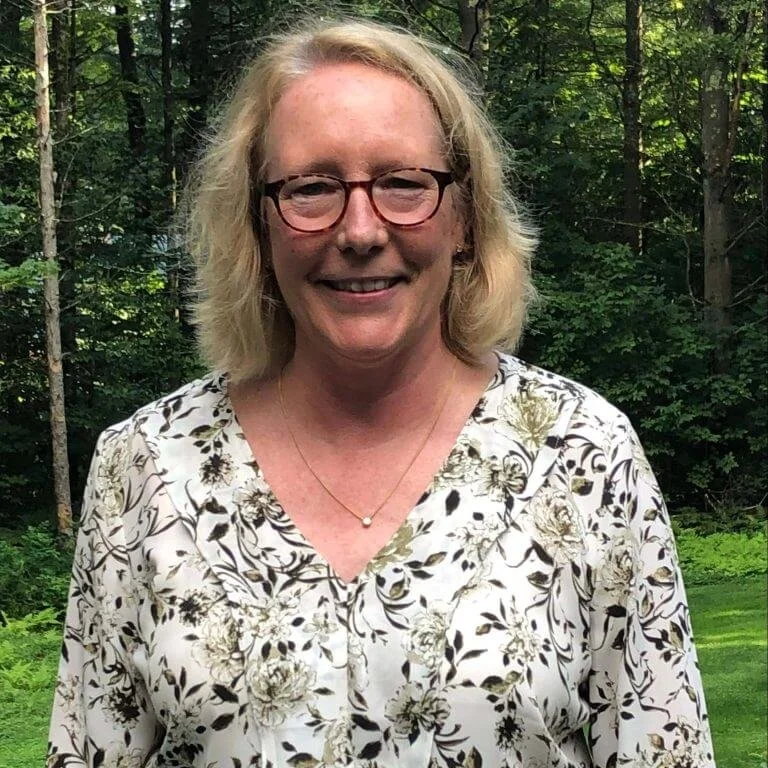 A woman with shoulder-length blonde hair, wearing glasses and a floral blouse, standing outdoors in a wooded area.