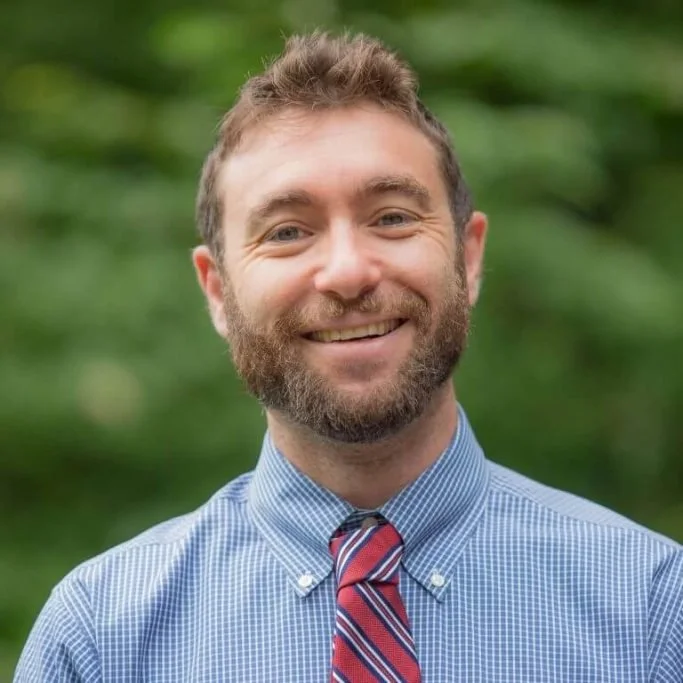A smiling man with a beard and short hair, wearing a blue checkered shirt and a striped red, white, and blue tie, standing outdoors with a blurred green background.