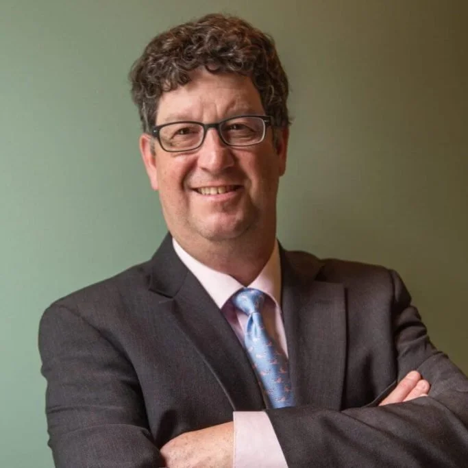 Portrait of a man in a suit with crossed arms, smiling, wearing glasses, against a plain green background.