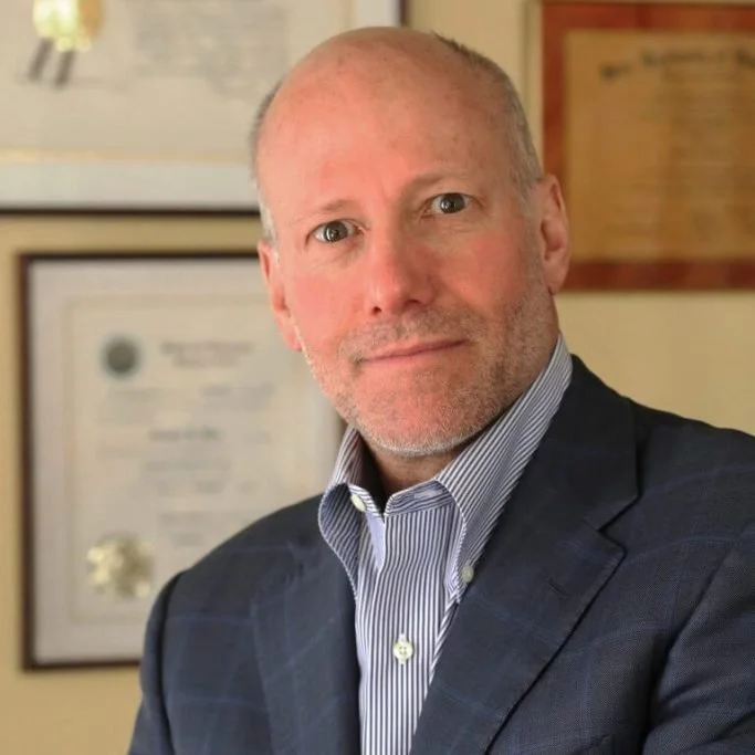 A middle-aged man with a bald head and stubble beard, wearing a dark suit and striped dress shirt, standing indoors in front of framed certificates and awards on the wall.