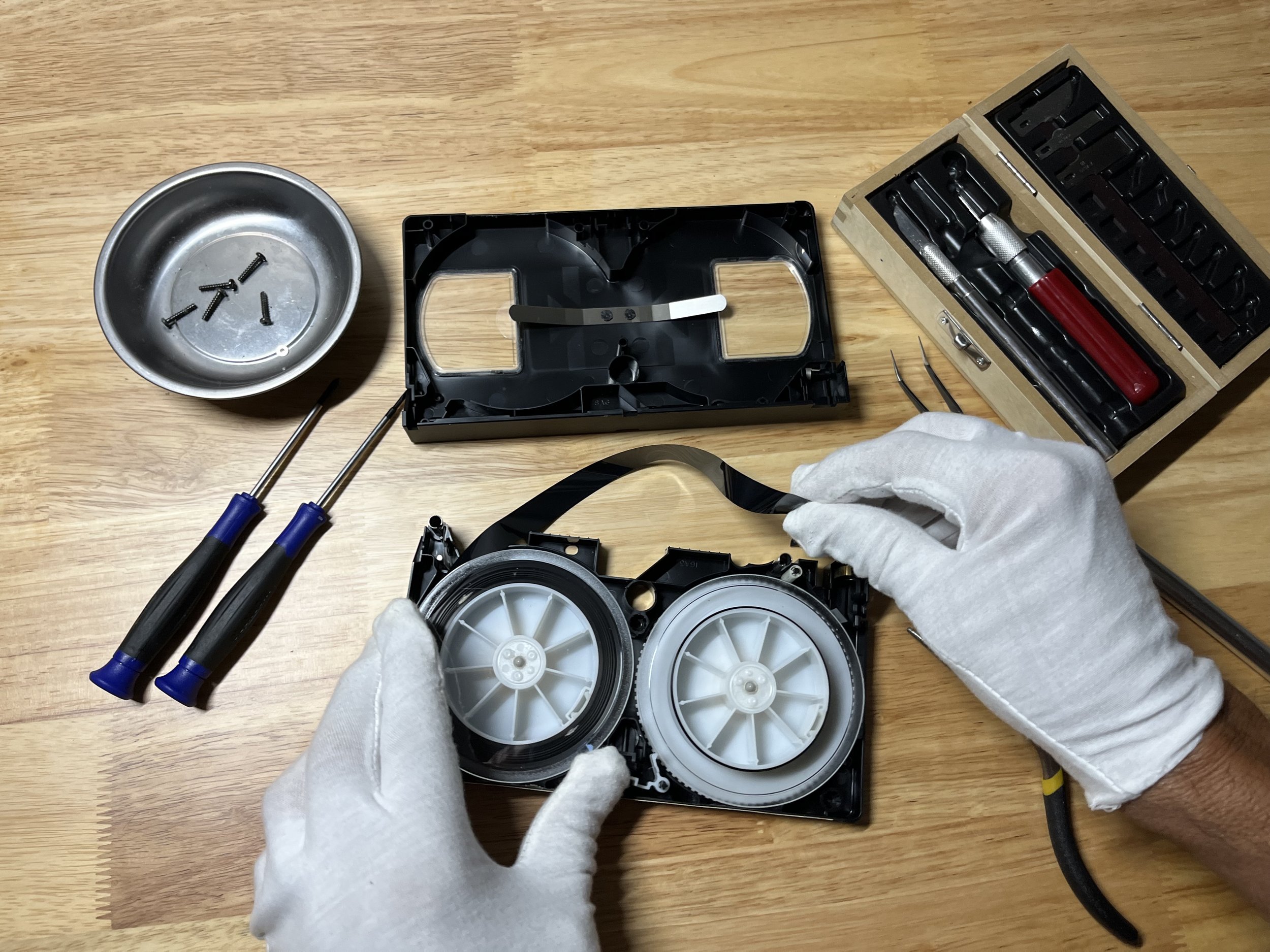 Hands in white gloves assembling a small electronic device with gears, surrounded by screwdrivers, a wooden box with tools, and a metal dish with screws on a wooden workbench.
