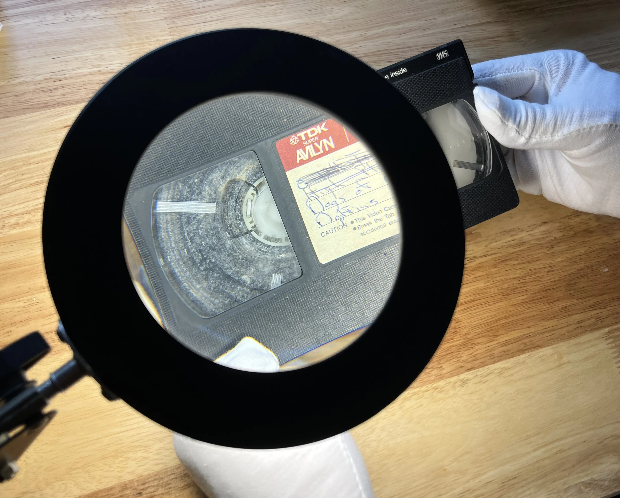 A person wearing a white glove holds an old VHS tape under a magnifying glass, revealing handwritten notes on the label.