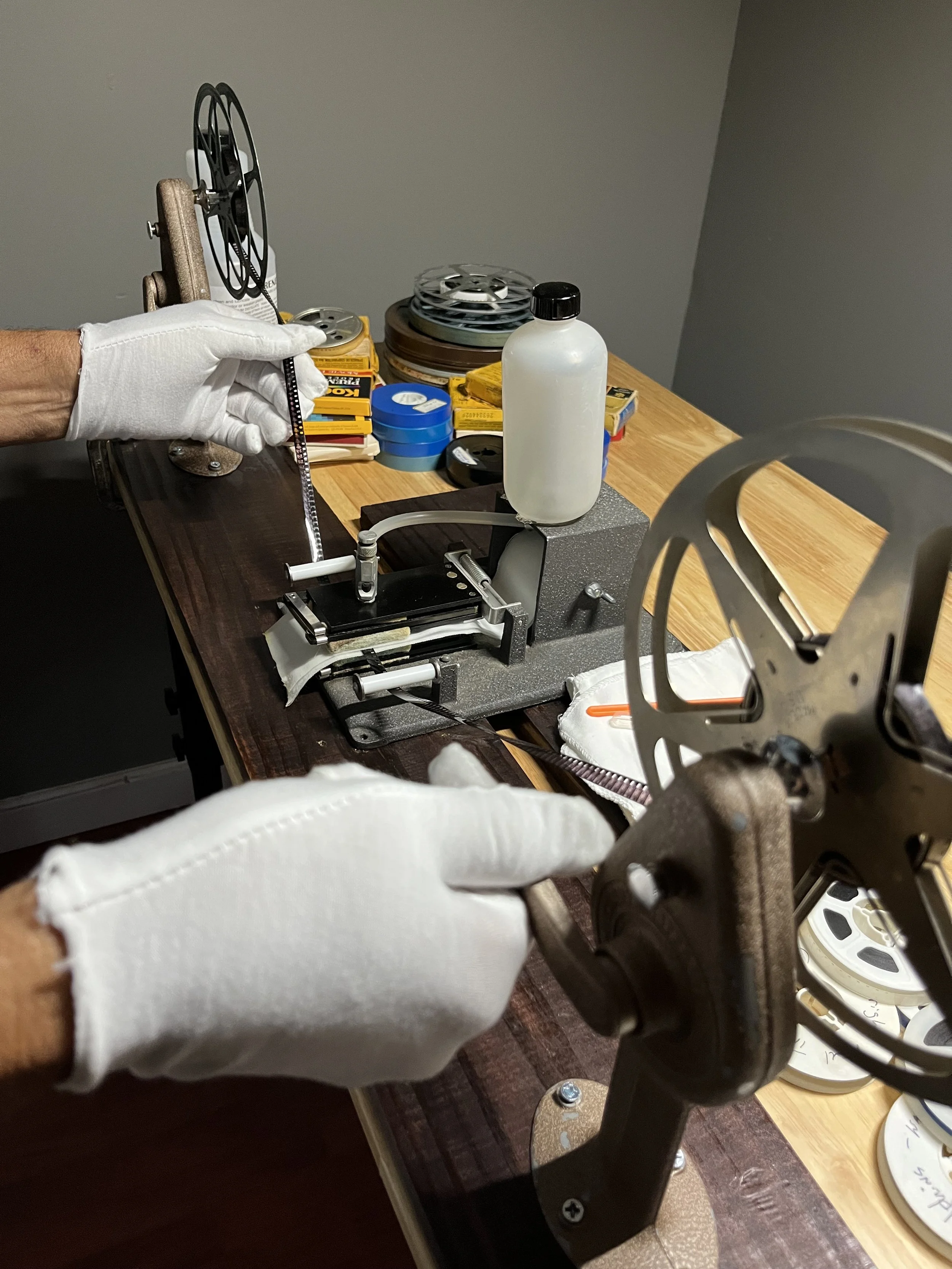 A person wearing white gloves working on a film reel in a film projection workshop, with various film and repair tools arranged on a wooden table.