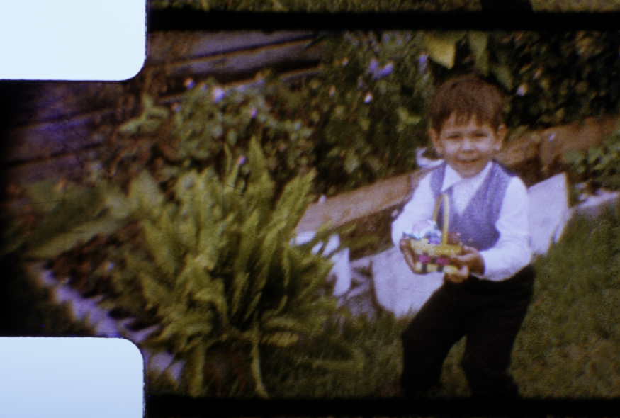 Young boy smiling and holding a basket of toys outdoors near a fence and bushes.