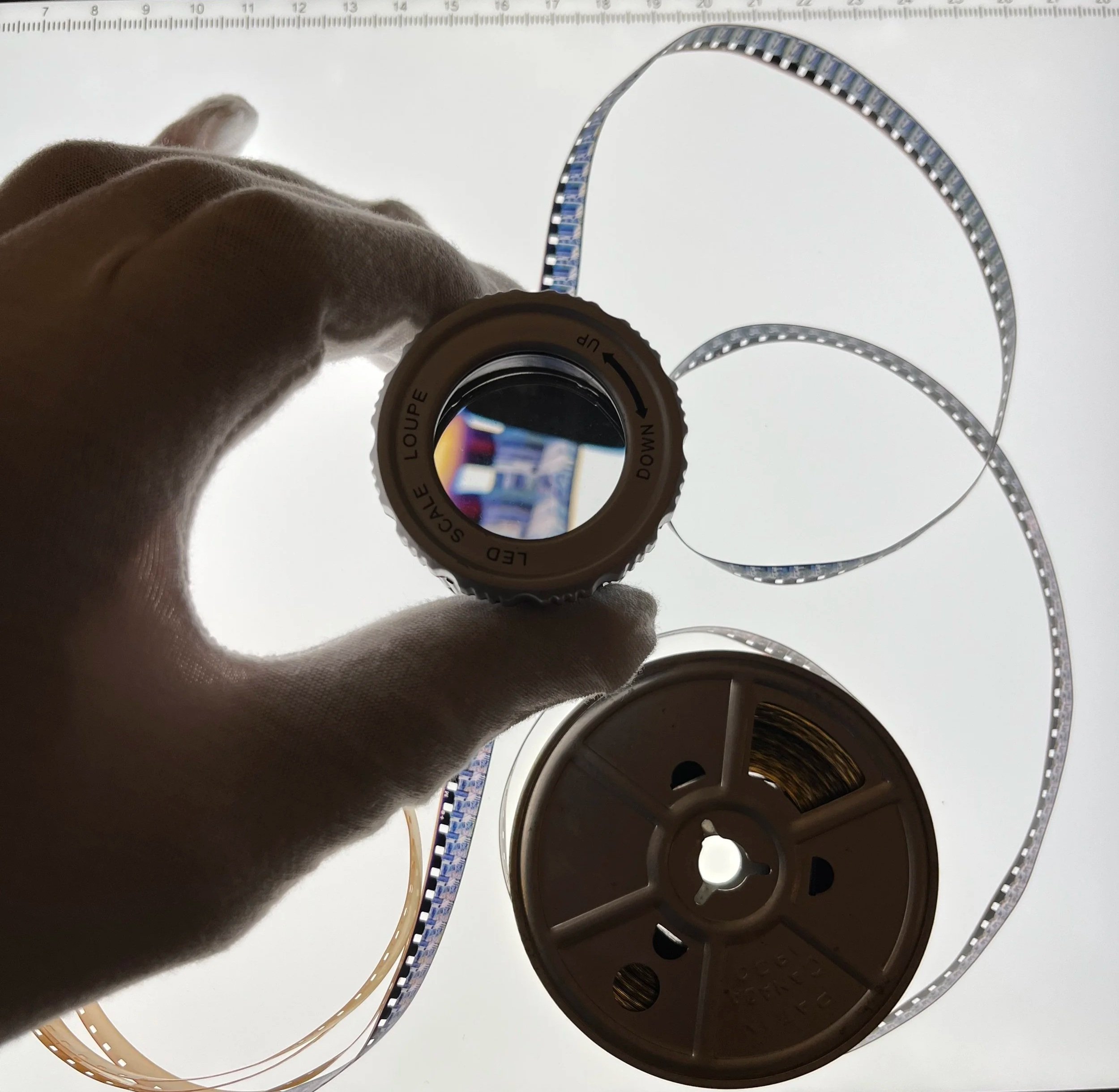A person's hand holding a small cylindrical lens with a labeled rotating ring, against a backlit white surface. Two reels of LED strip lights and a flexible LED strip are also visible.