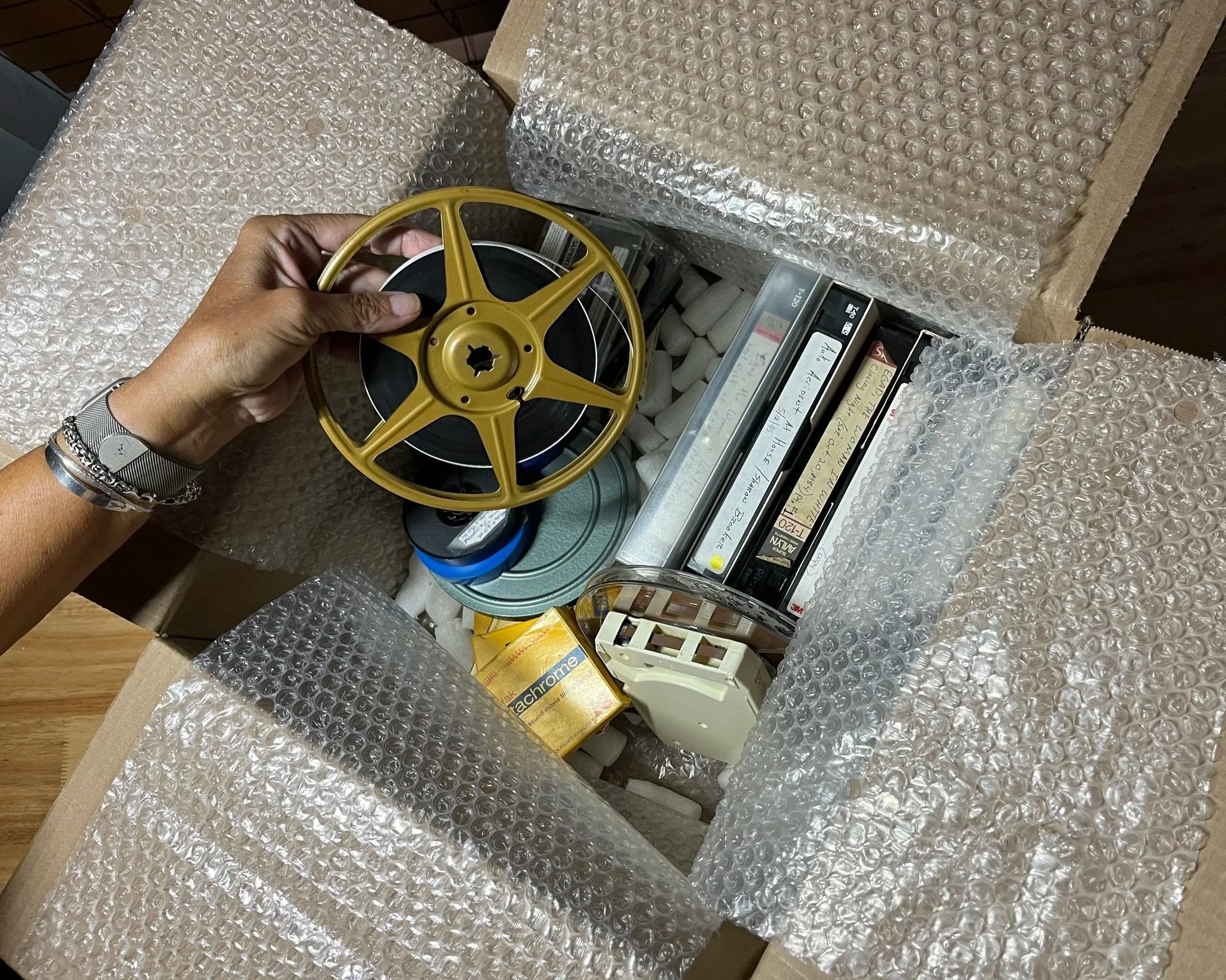Person unboxing a cardboard box filled with vintage audio and video tapes, a film reel, and other retro electronic items, surrounded by bubble wrap.