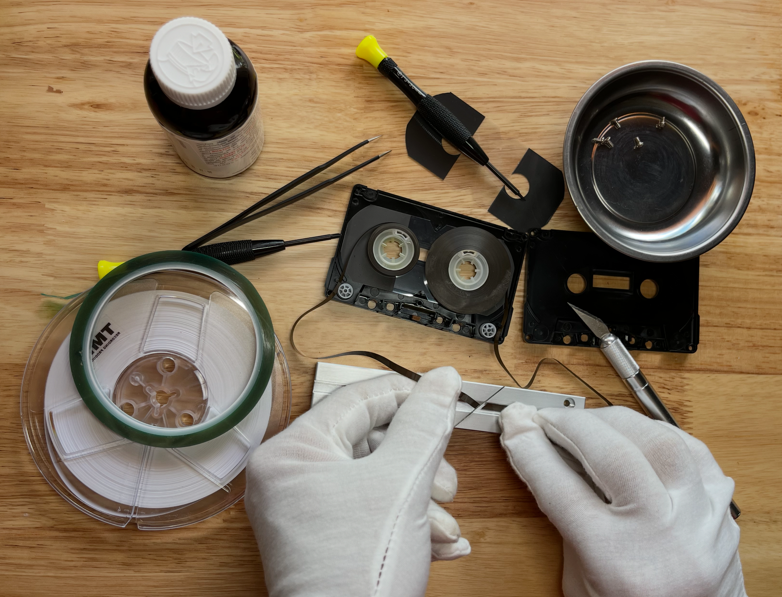 Person wearing white gloves working with cassette tapes, tools, and equipment on a wooden surface, including a screwdriver and a bottle of cleaning solution.