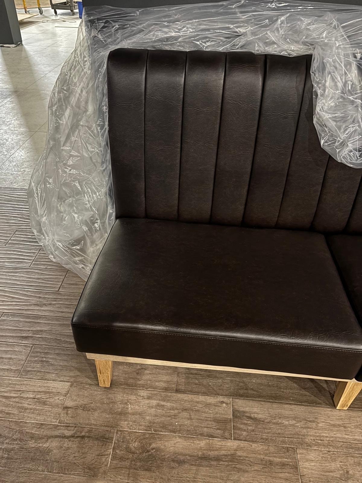A black upholstered chair with wooden legs, wrapped in clear plastic, placed on a wooden floor in a room with gray tiles.