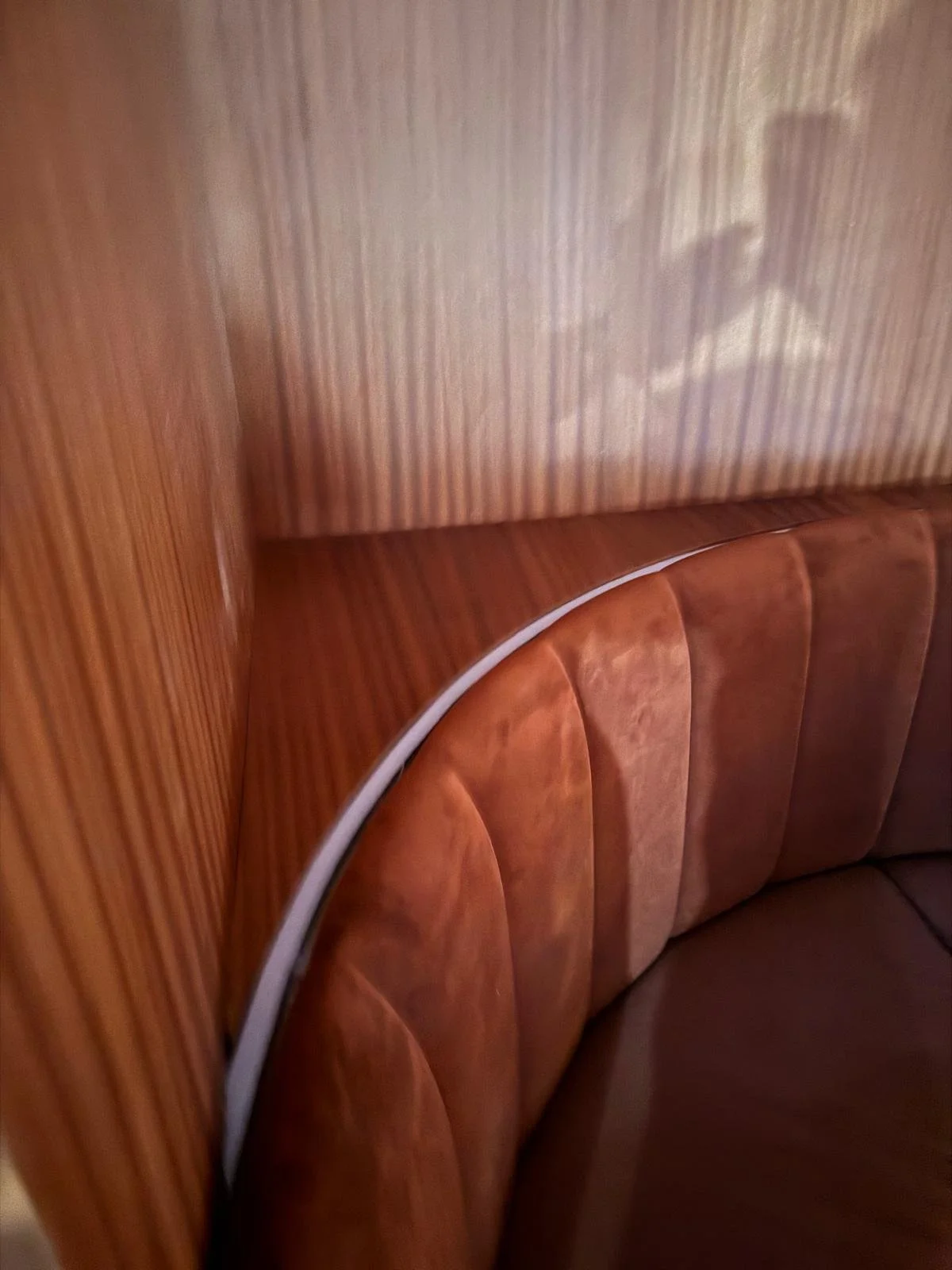 Close-up of a vintage wooden booth seat with a rounded, padded backrest and a white trim along the edge, reflected in the textured wall.