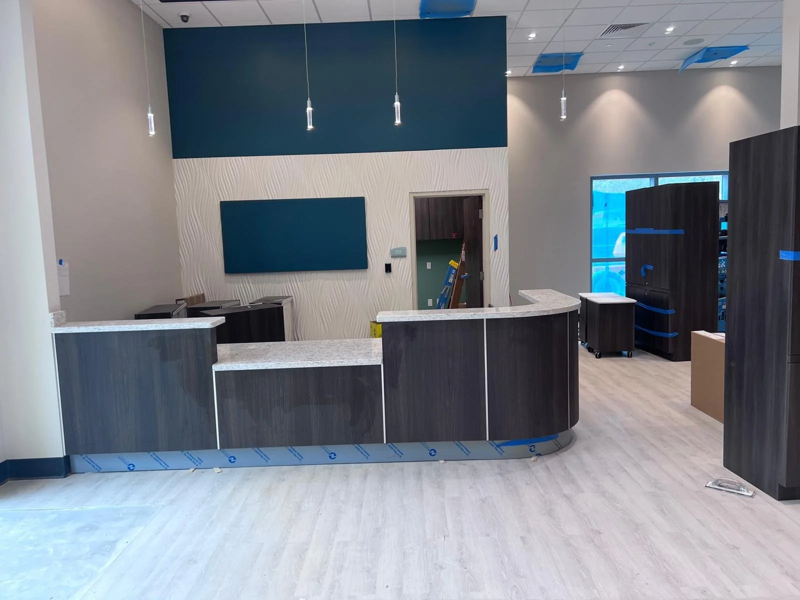Interior of a modern office reception or front desk area with dark wood and marble surfaces, a blue accent wall, pendant lighting, a large window, and partially installed ceiling panels.