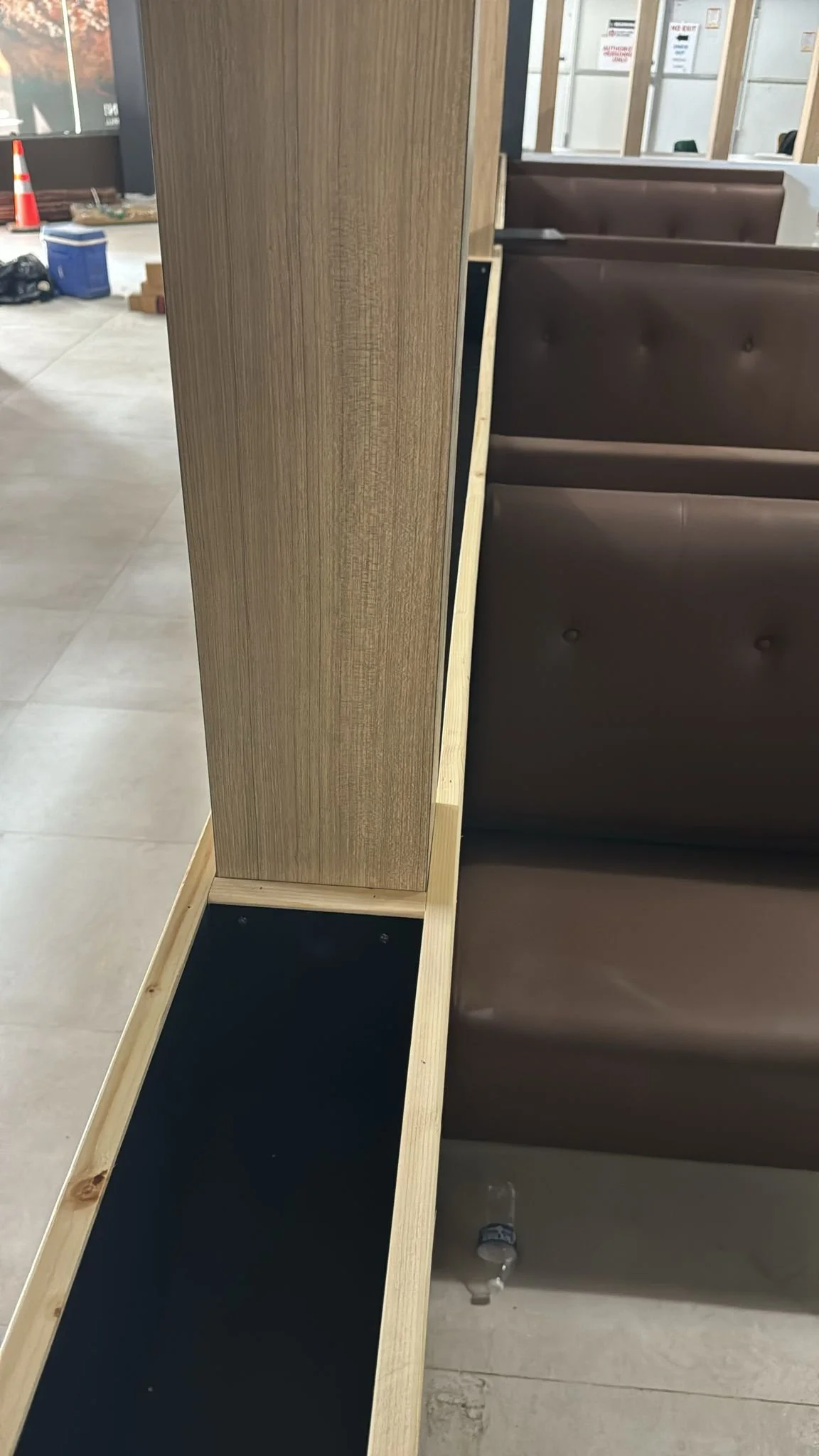 Wooden booth divider with a black panel at the bottom, situated next to a brown upholstered booth seat inside a fast-food restaurant under construction.