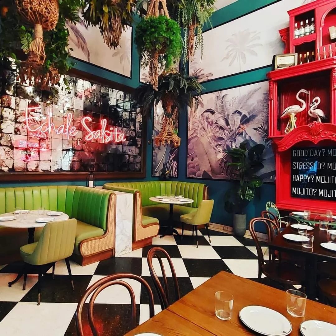 Interior of a restaurant with black and white checkered floor, green booth seating, wooden tables, and decorative tropical wall murals. There is a neon sign reading 'Chale Salsa' and a red cabinet with flamingo decor.
