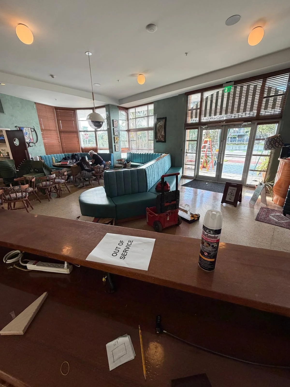 Inside a cafe or restaurant with green walls, large windows, and a mix of wooden and upholstered furniture. Two people sit at a table near the windows. An 'Out of Service' sign is on the front desk, where a can of spray is also visible.