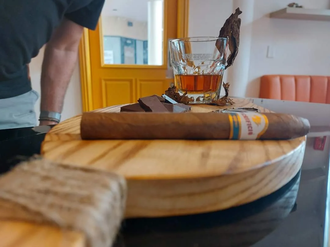 A wooden serving board with a cigar, a glass of whiskey, and chunks of dark chocolate on it, on a dark surface. In the background, a person wearing a black shirt and gray pants is leaning over a table, with yellow door and windows behind.