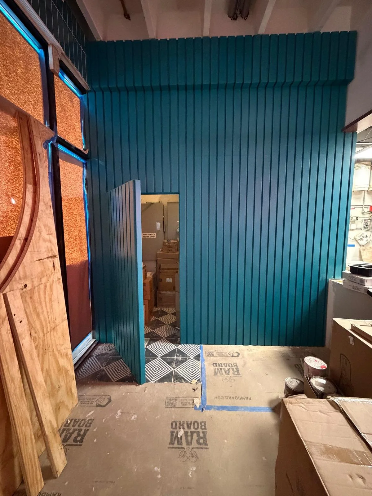 Interior of a room under construction with a partially installed turquoise slatted wall and door frame, cardboard boxes, paint cans, and exposed ceiling beams.