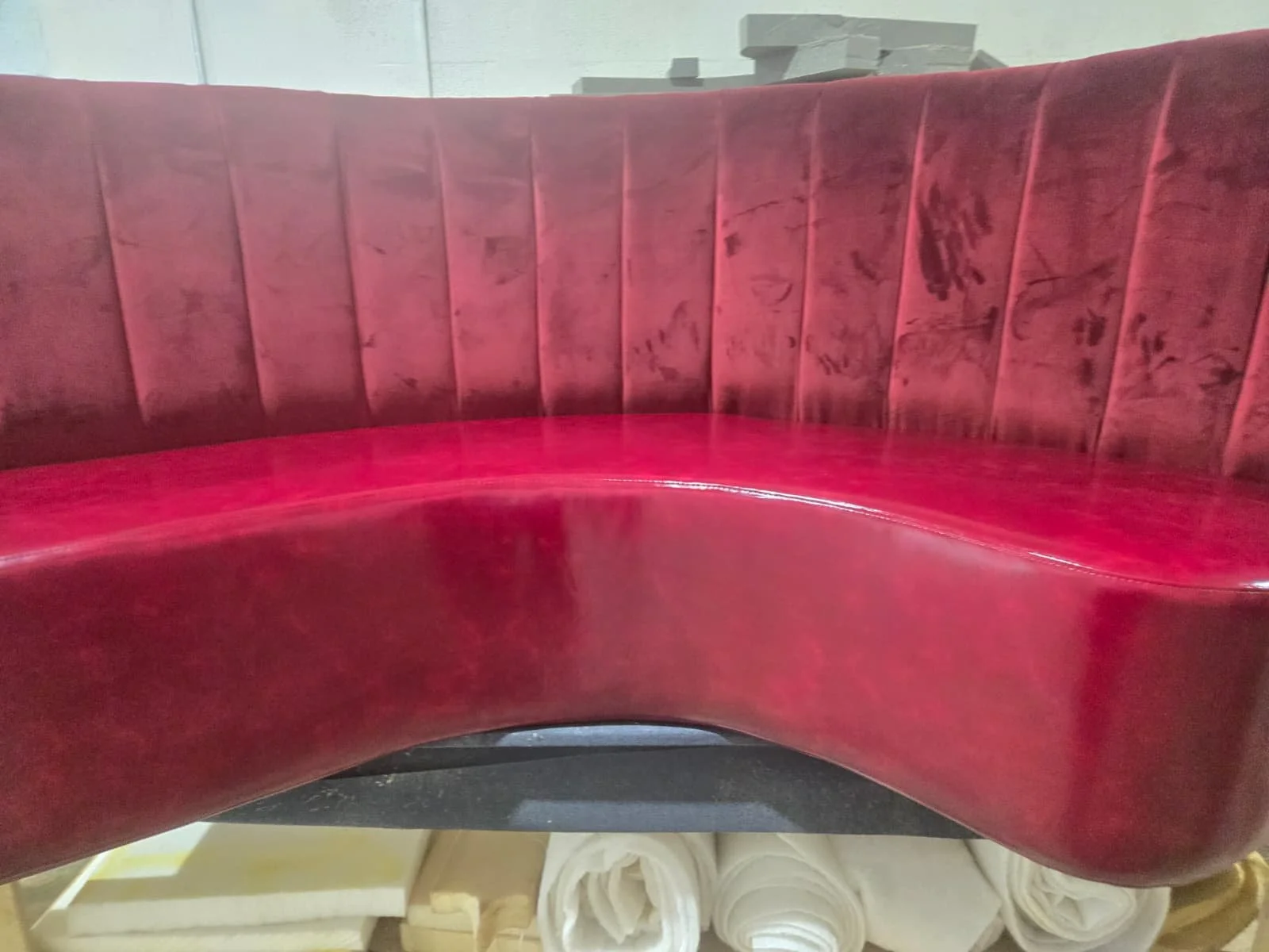 Red upholstered curved booth seating with vertical stitching on the backrest and visible scratches on the seat cushion, with a stack of white rolled fabrics or towels in the background.