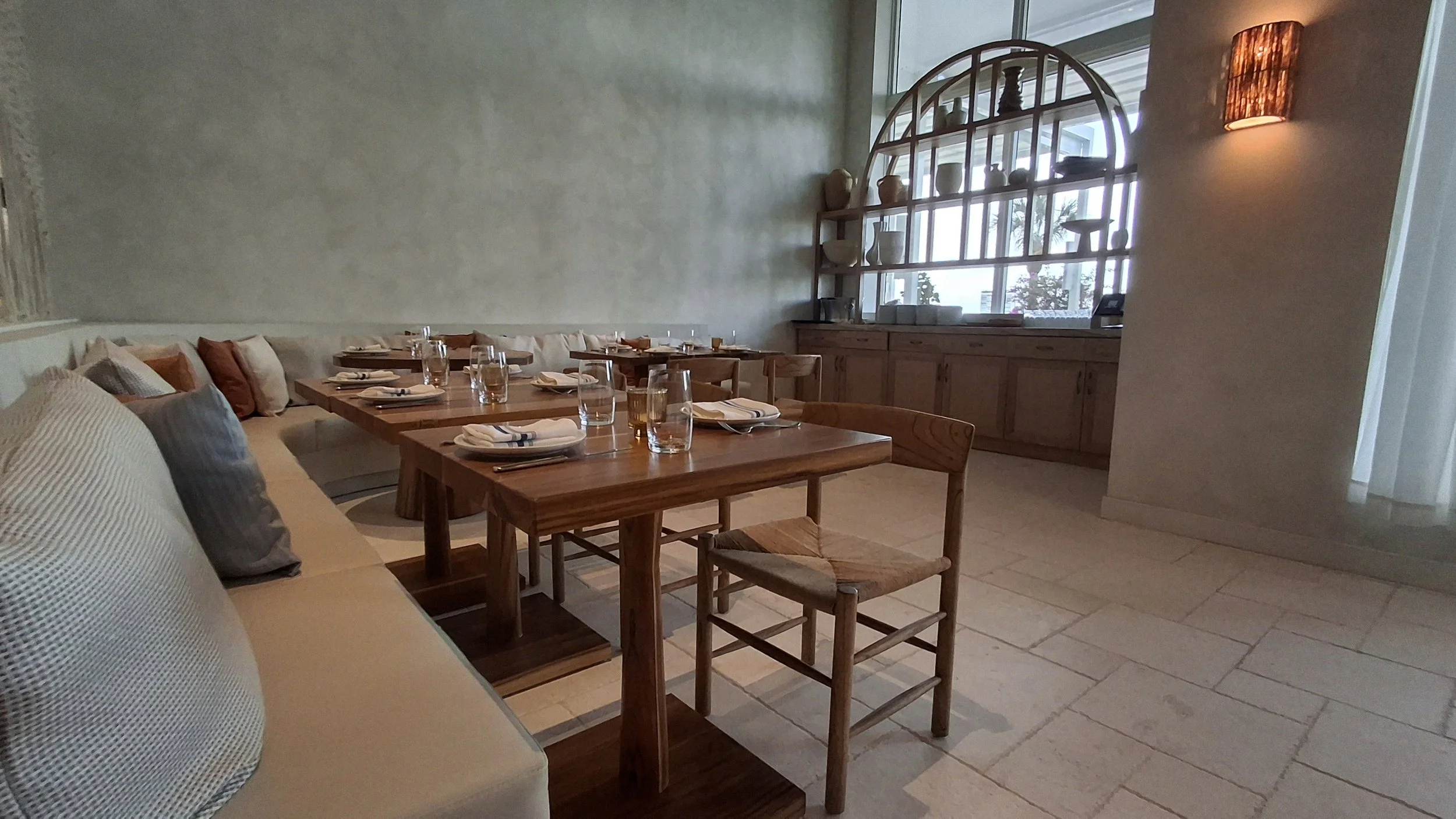 A dining area with wooden tables set with glasses, napkins, and utensils; a cushioned seating area with pillows; a large window with shelves displaying pottery; warm lighting on the wall.