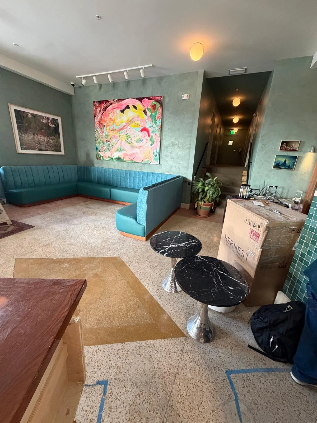 A living room with a teal sectional sofa, colorful abstract paintings on green walls, and two black marble side tables. There are staircase and hallway in the background, a potted plant, and a large cardboard box labeled 'HerneSt' near the wall.