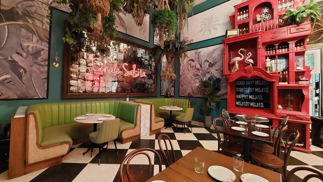 Interior of a restaurant with green booth seating, round white tables, and wooden chairs on a black-and-white checkered floor. There is a large red hutch with various jars and decorations, a blackboard with a menu, and tropical-themed artwork on the 