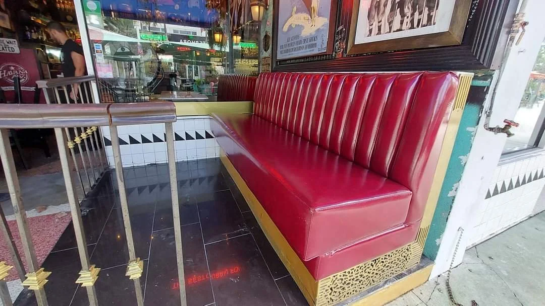 Red booth seat with yellow trim in a restaurant or cafe. Part of a wine-themed decor with wine posters on the wall, near a window and a sidewalk outside.