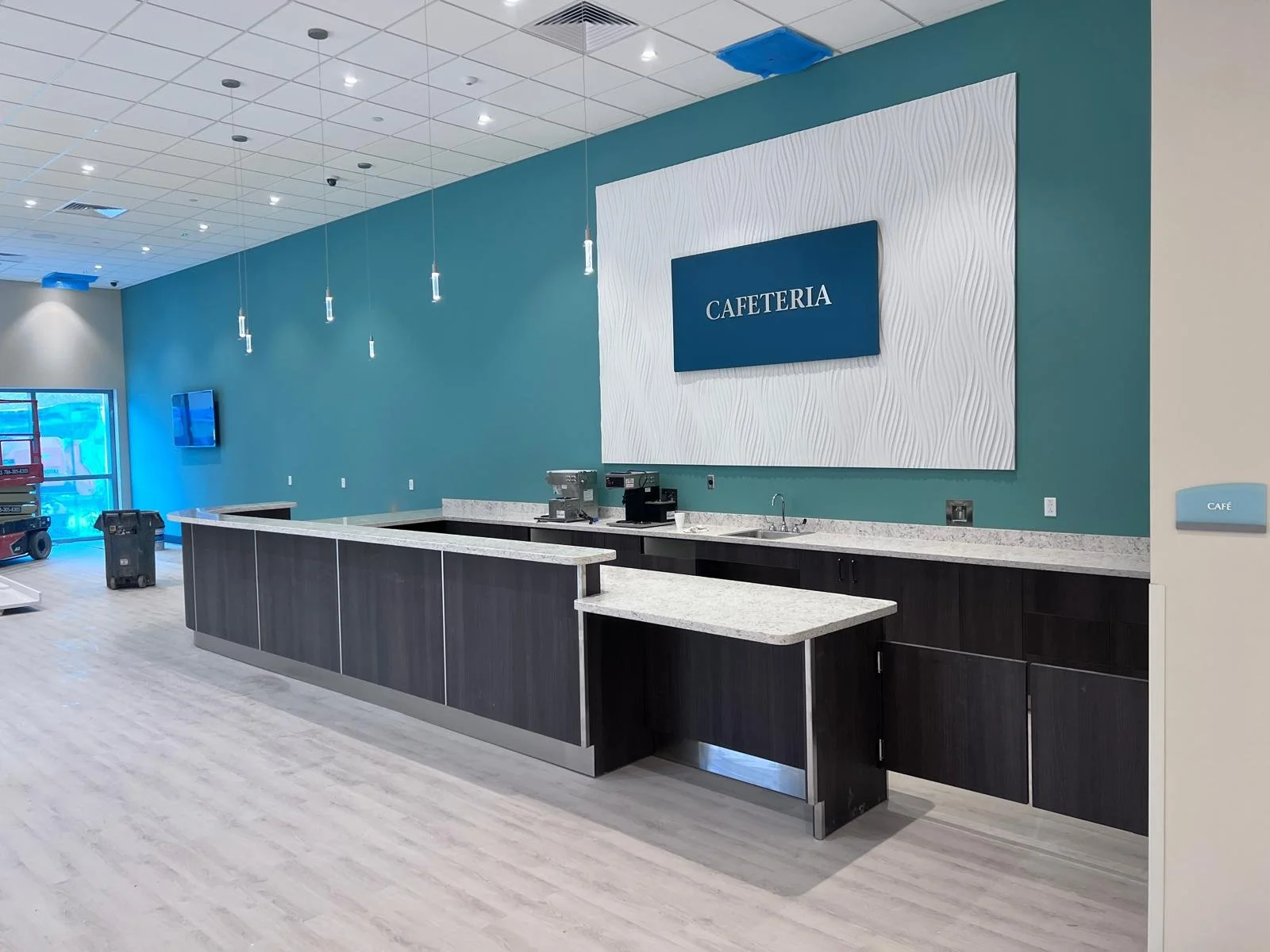 Empty cafeteria counter with coffee machines, a sink, and a sign that says 'CAFETERIA' on a blue wall, with hanging lights and a TV in the background.