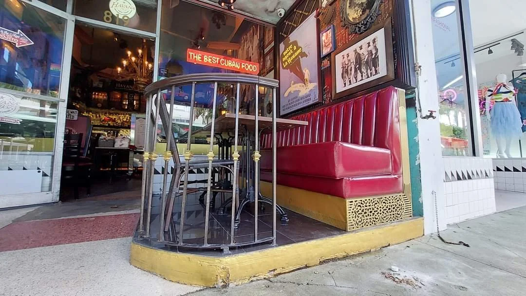 Street view of a Cuban restaurant with a red booth, a high table with stacked chairs, framed pictures, and a neon sign that says "The Best Cuban Food."