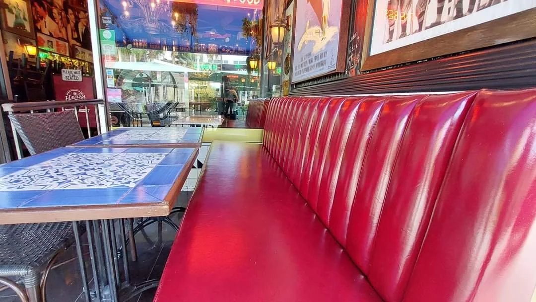 Empty restaurant booth with a long red leather seat and a wooden table with blue and white tiles, outside view through glass windows showing people walking on the street.