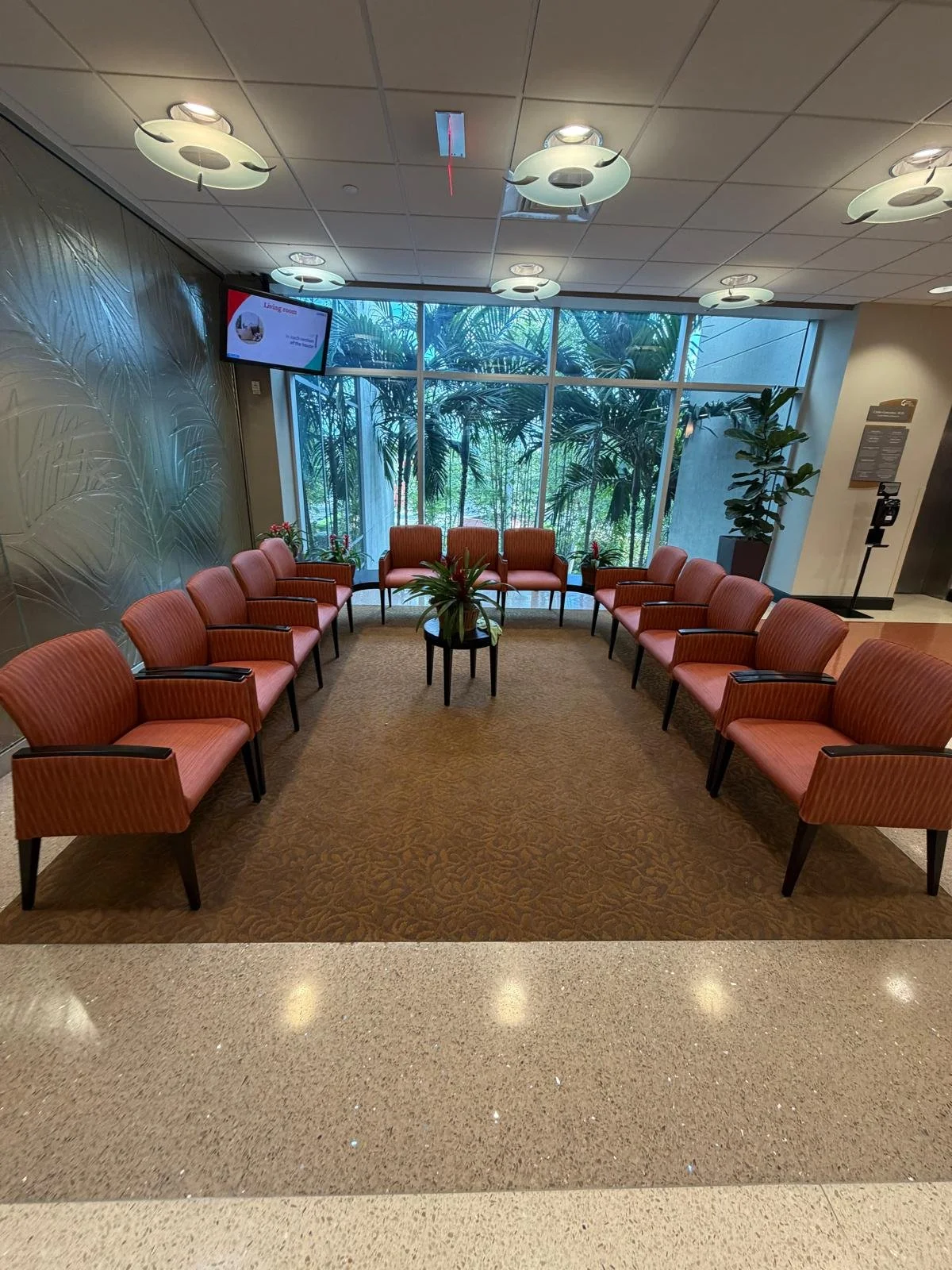 Waiting room with 14 pink upholstered chairs arranged in a circle around a small black table with a potted plant, large window with tropical plants outside, ceiling lights, wall with textured palm leaf pattern, flat-screen TV, and a potted plant near