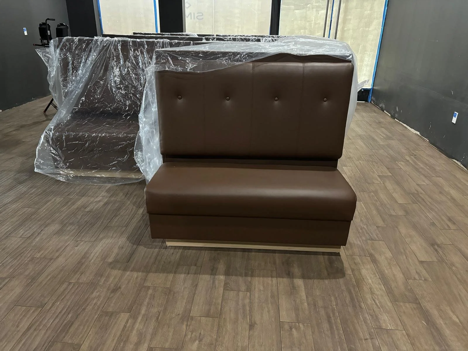Brown booth seats, one with a backrest and one without, wrapped in plastic, in a room with wood-like floor and dark walls.