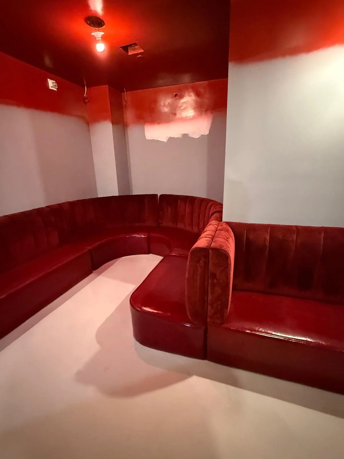 A red corner booth with velvet upholstery in a dimly lit room with red lighting and a plain white floor.