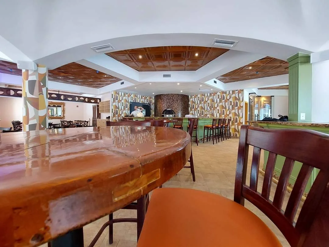 Interior of a restaurant with wooden tables and chairs, a bar with bar stools, patterned wallpaper, and a pizza oven in the background.