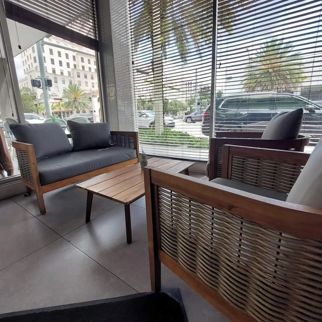 Interior of a cafe with wooden furniture, including a two-seater sofa, two chairs, and a small coffee table, in front of large windows with horizontal blinds showing palm trees and a city street outside.