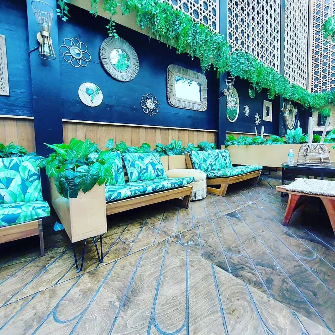 Indoor seating area with wooden floor, green plants, patterned cushions, and decorative wall artwork including mirrors and flower shapes.