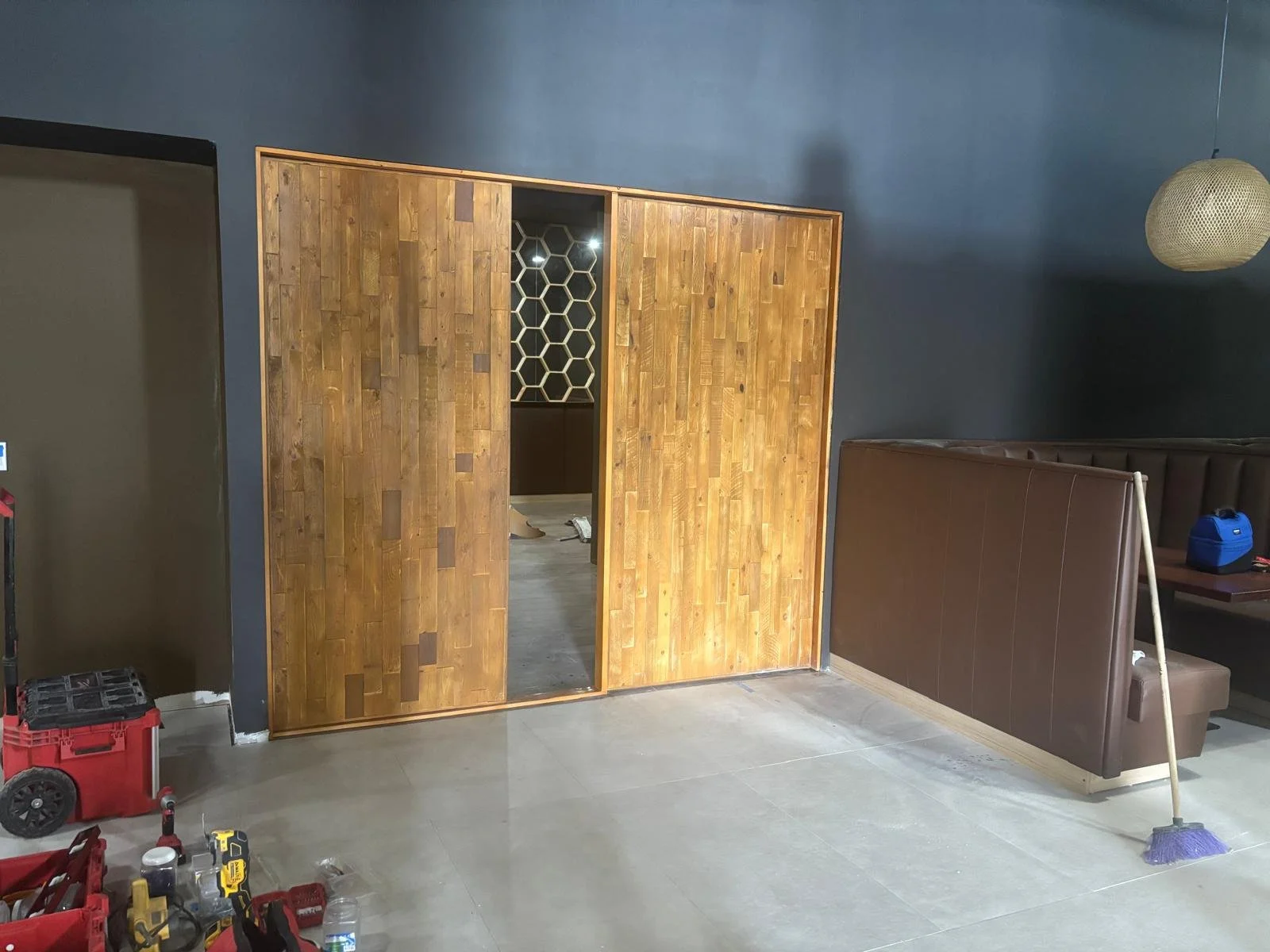 Interior of a room under construction with a wooden sliding door, dark gray walls, seating booth, tools, and cleaning supplies