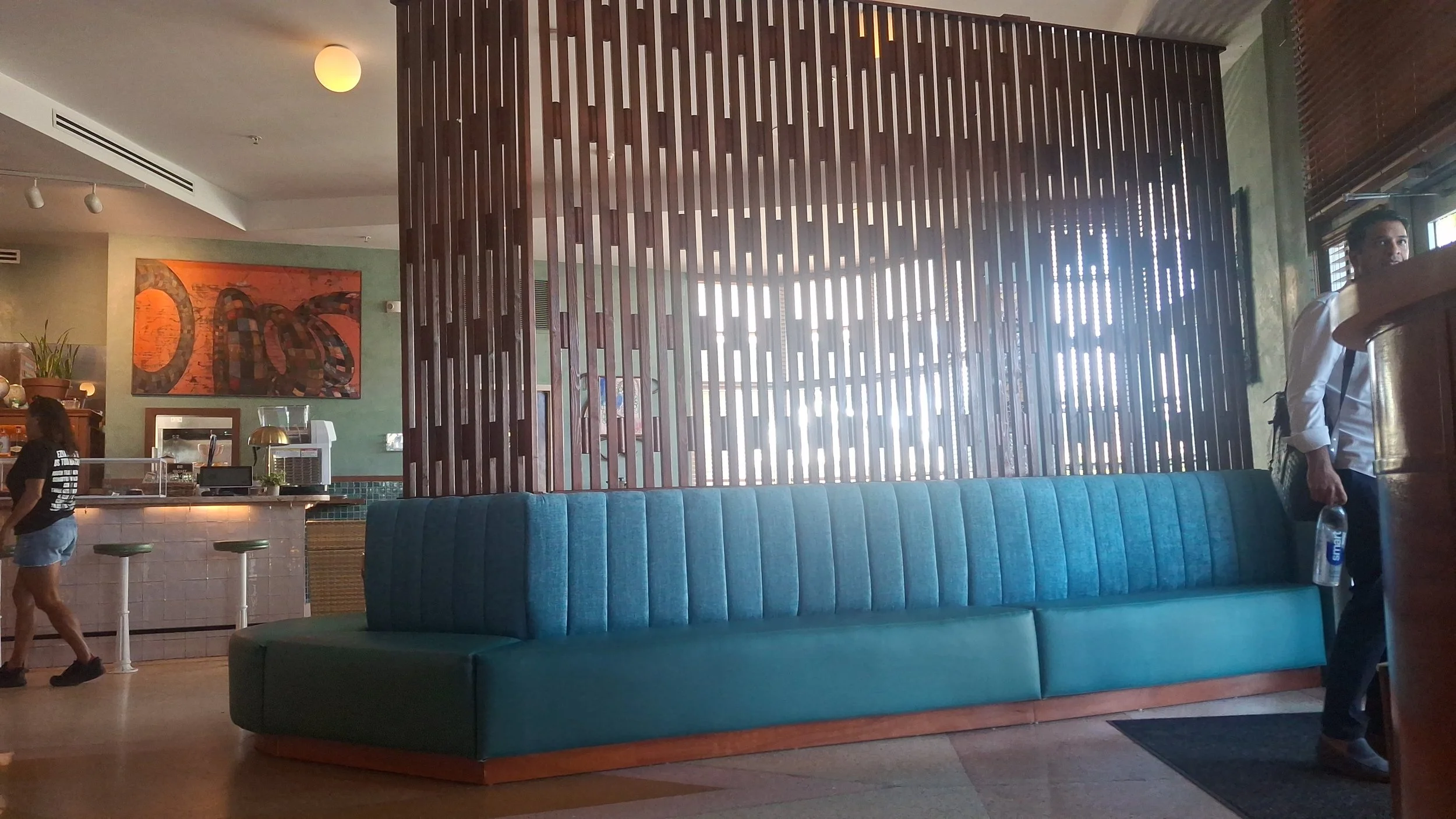 Interior of a restaurant or cafe featuring a blue upholstered booth, a wooden slat partition, a bar area with white tiles and bar stools, and artwork on the green wall. There are a few people visible, one woman walking and a man near the entrance.