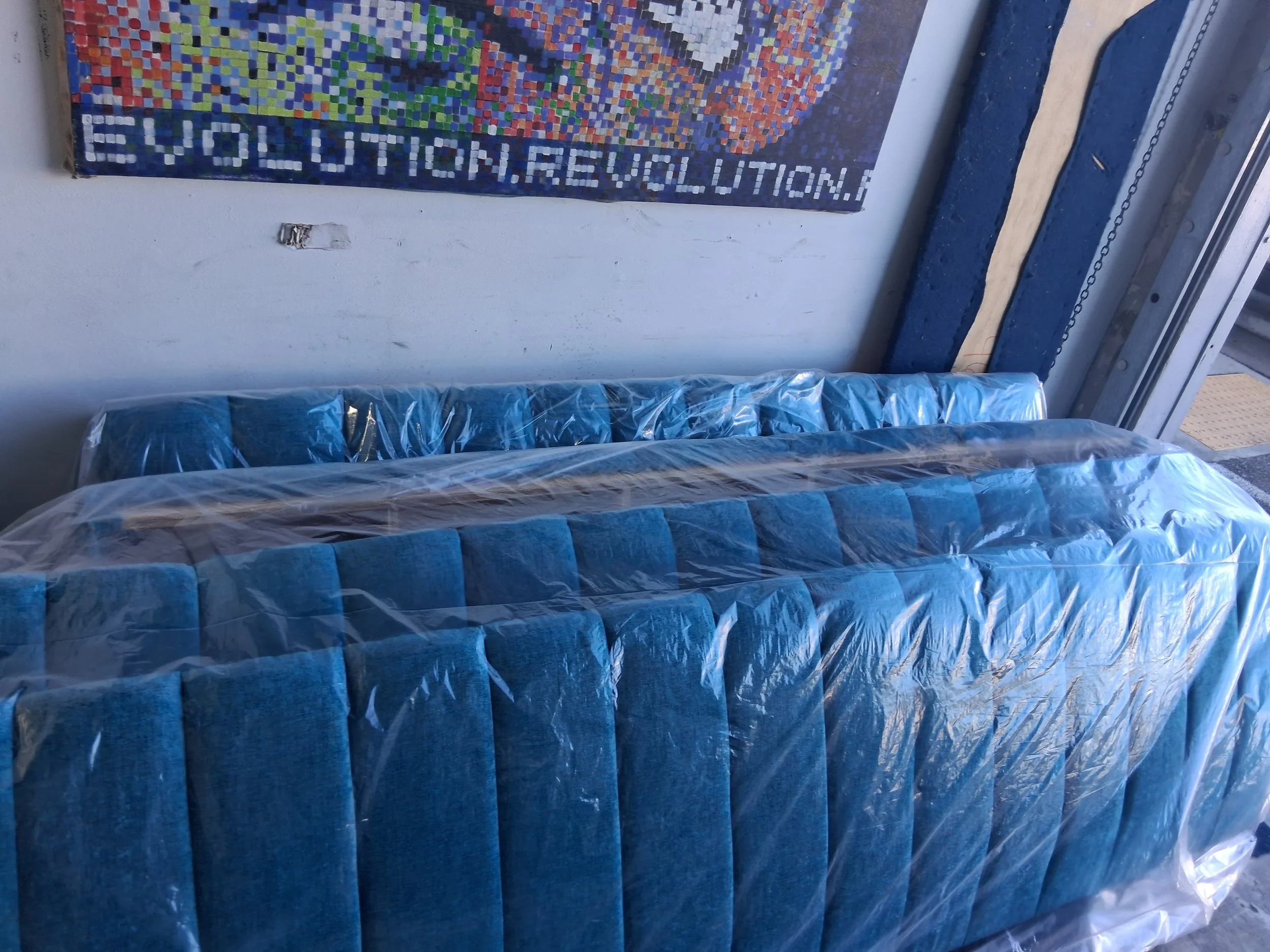View of wrapped mattresses in plastic in a warehouse or storage area, with a colorful mosaic artwork and partially visible elevator or structure nearby.