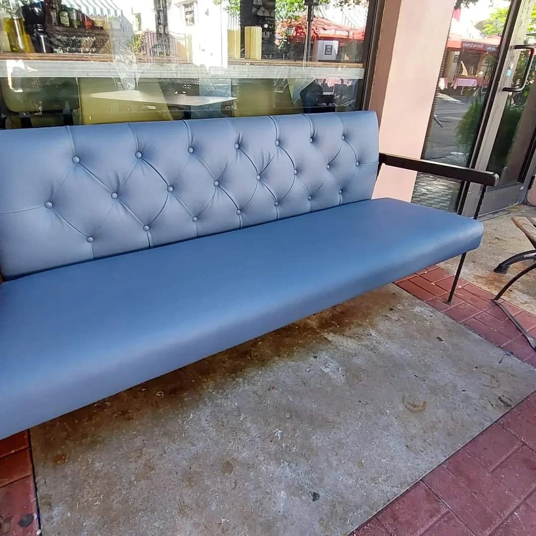 Blue padded bench with button tufting, armrest on the right side, outside on a sidewalk next to a glass door and window. Reflections of outdoor tables and umbrellas can be seen in the glass.