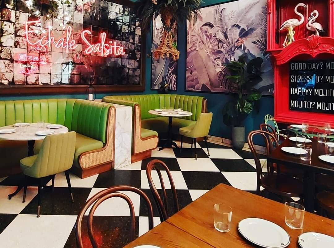 Inside a vibrant restaurant with green booths, wooden chairs, checkered black and white floor, and tropical decor including flamingos and palm leaves.