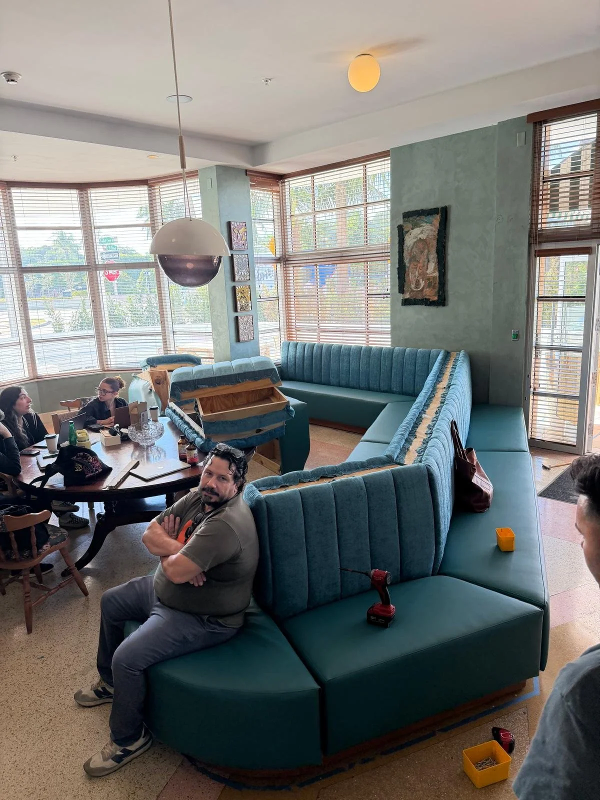 Interior of a restaurant or cafe with large windows, turquoise blue booth seating, and a group of people sitting at a table. A man sits in the foreground with arms crossed, and several others are engaged in conversation. There are tools and supplies,