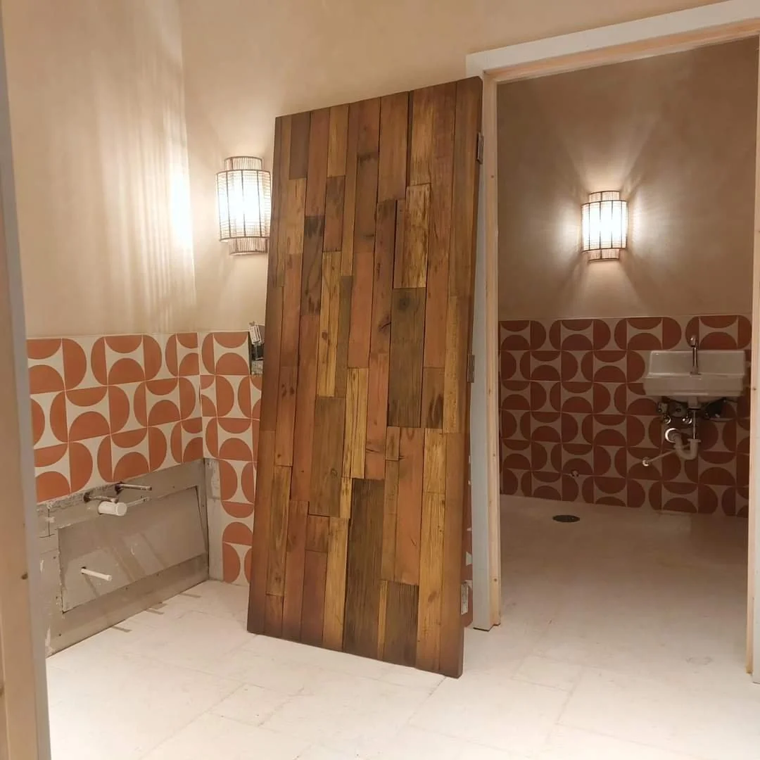 Interior of a room with retro orange patterned wall tiles, a wooden sliding door, a small white sink, and two wall-mounted light fixtures.