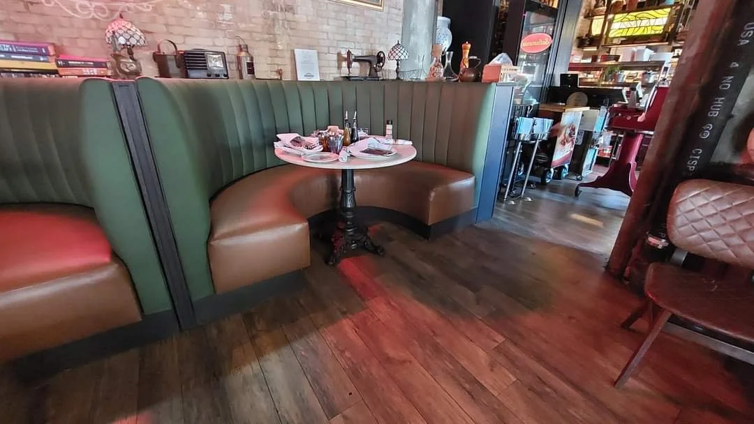 A cozy restaurant booth with green and brown leather seating, set with plates, glasses, and cutlery on a small round table. The background shows a brick wall with decorative lamps and a shelf filled with books and decorative items. The floor is woode