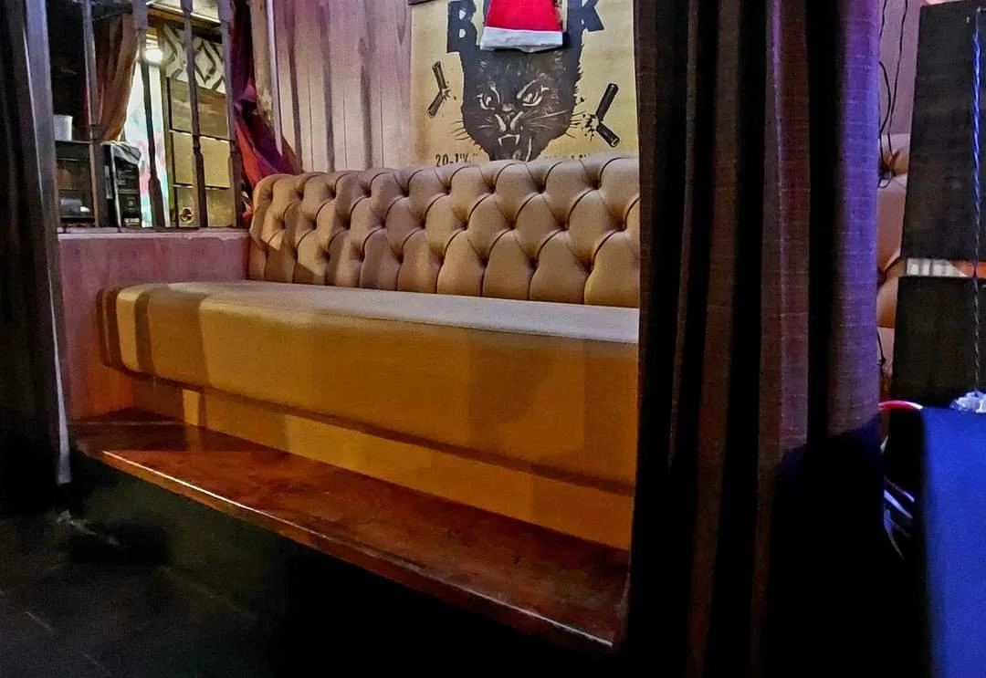 A beige tufted leather couch in a cozy, dimly lit room with a decorative wall featuring a large black panther poster or artwork, surrounded by various small objects and a table behind it.
