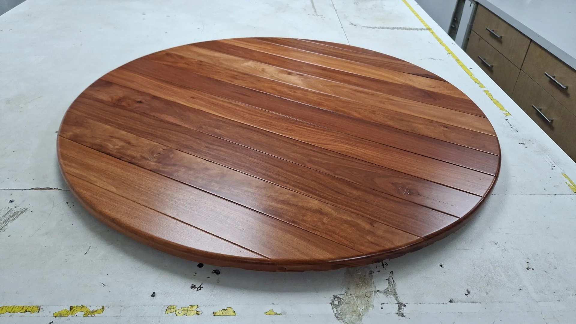A round wooden table with a polished surface made of horizontal wooden planks, placed on a workshop or fabrication area.