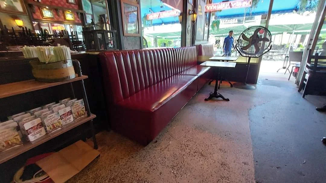 Red booth seating inside a restaurant next to a window, with a small table, a stand fan, and a shelf holding napkins and condiments.
